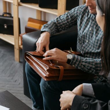 A man demonstrates a brown leather briefcase to a woman in a stylish showroom.