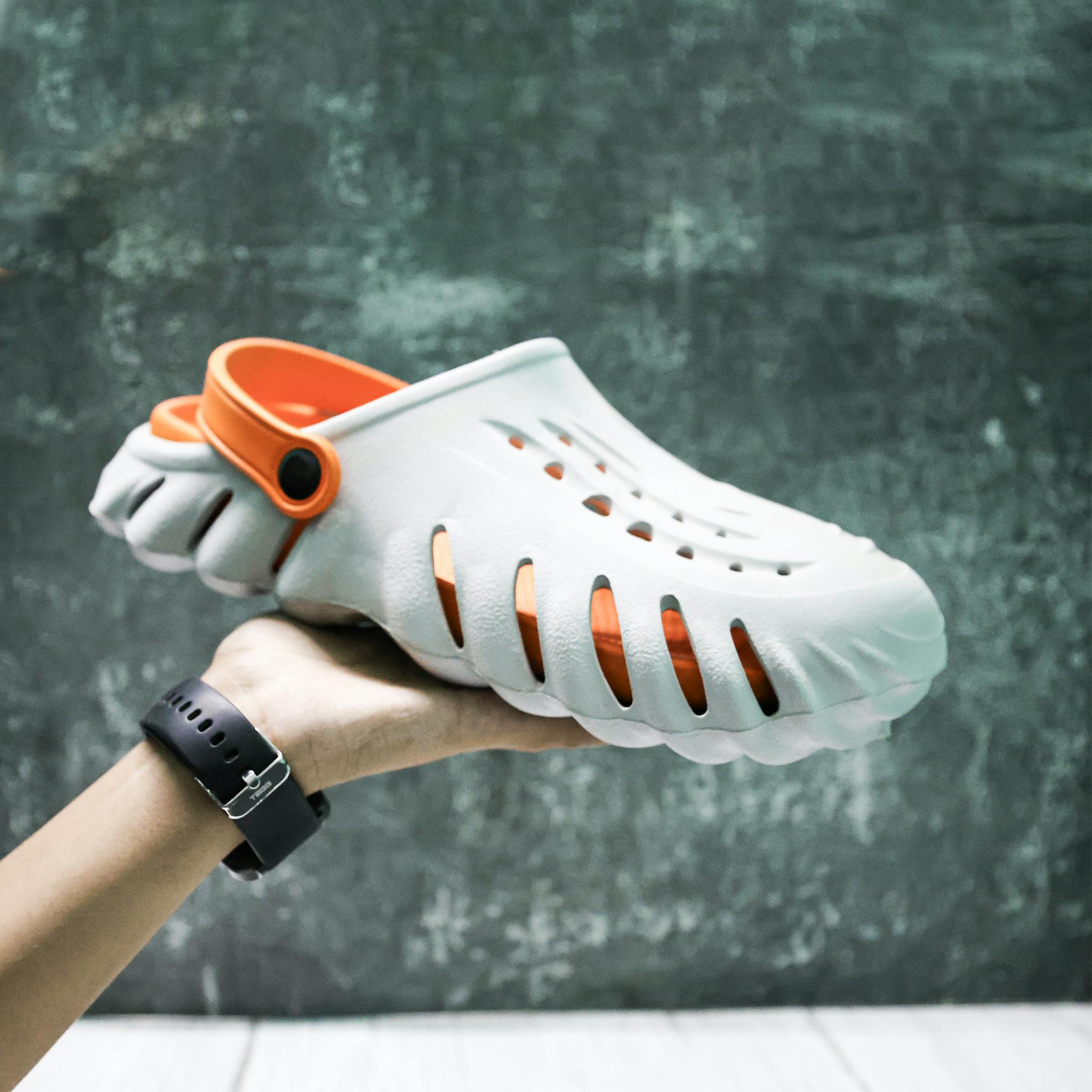 Close-up of a hand holding a stylish white and orange clog shoe against a textured background.